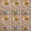 Set of six wedding-themed wine glass charms displayed on white wine glasses filled with pale white wine. Each charm features colorful beads and silver-tone pendants including Bride, Groom, heart, key, lock, and bow designs. Styled on a taupe linen tablecloth for an elegant wedding table setting.